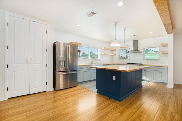 kitchen featuring a kitchen island, decorative light fixtures, stainless steel fridge with ice dispenser, wall chimney range hood, and light hardwood / wood-style flooring