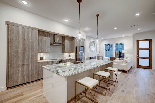 kitchen featuring sink, backsplash, hanging light fixtures, wall chimney range hood, and a center island with sink