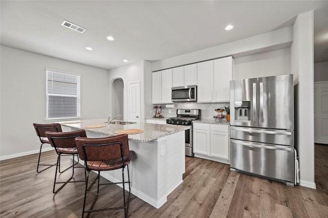 kitchen featuring appliances with stainless steel finishes, light stone countertops, a kitchen island with sink, decorative backsplash, and white cabinets