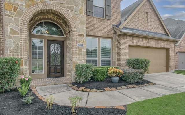 doorway to property featuring brick siding, a shingled roof, an attached garage, stone siding, and driveway