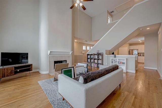 living room featuring ceiling fan, a towering ceiling, a tiled fireplace, and light wood-type flooring