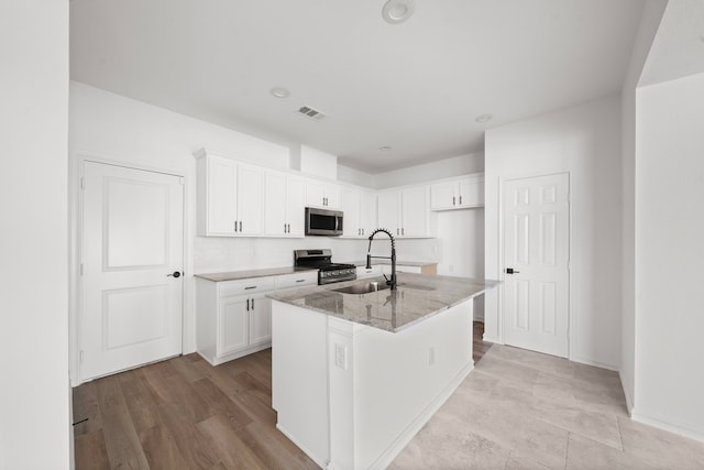 kitchen with sink, a kitchen island with sink, white cabinetry, stainless steel appliances, and light stone counters