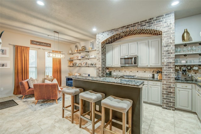 kitchen featuring appliances with stainless steel finishes, decorative light fixtures, tasteful backsplash, dark stone counters, and a center island with sink