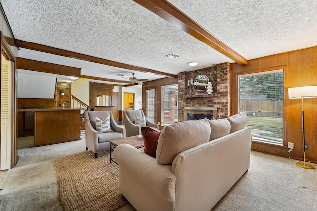 carpeted living room featuring a brick fireplace, a textured ceiling, wooden walls, beamed ceiling, and ceiling fan