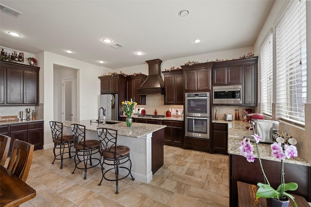 kitchen with sink, custom exhaust hood, an island with sink, stainless steel appliances, and light stone countertops