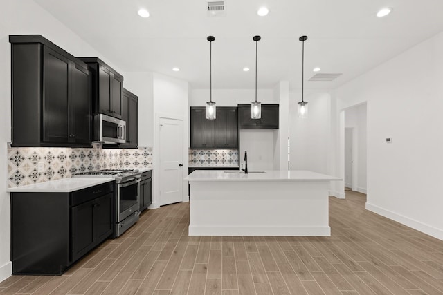 kitchen with sink, appliances with stainless steel finishes, pendant lighting, a kitchen island with sink, and decorative backsplash