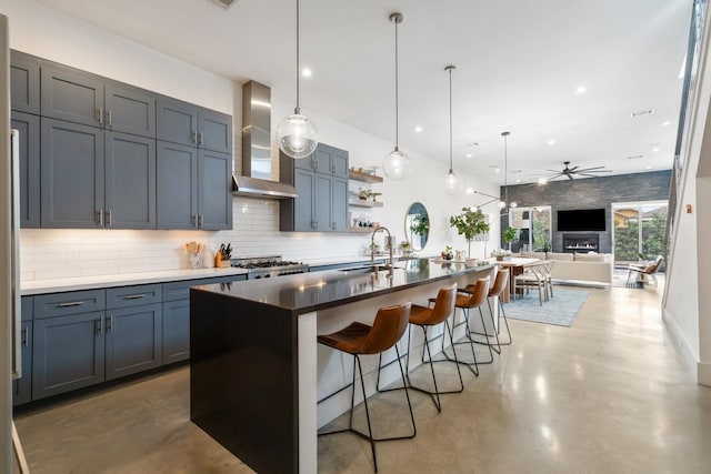 kitchen featuring pendant lighting, wall chimney range hood, a breakfast bar, ceiling fan, and an island with sink