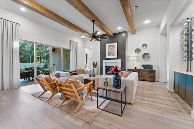 living room featuring a tiled fireplace, ceiling fan, light wood-type flooring, and beamed ceiling