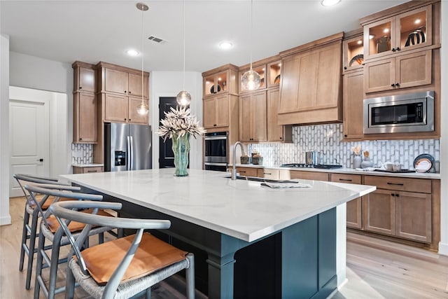 kitchen featuring pendant lighting, stainless steel appliances, light stone countertops, a center island with sink, and light hardwood / wood-style flooring