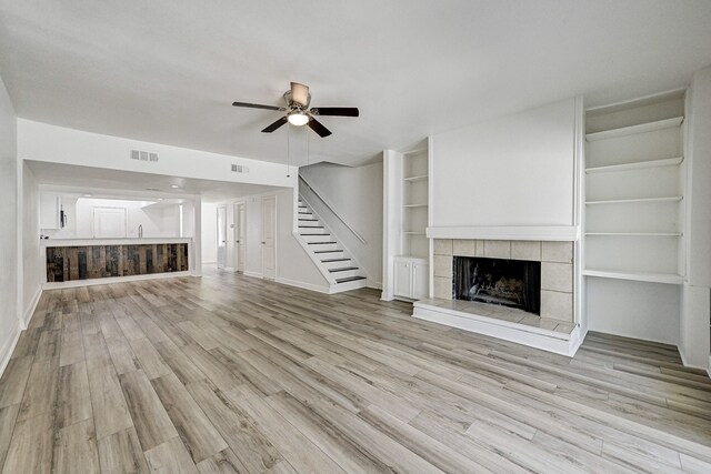 unfurnished living room featuring a tiled fireplace, built in shelves, ceiling fan, and light wood-type flooring