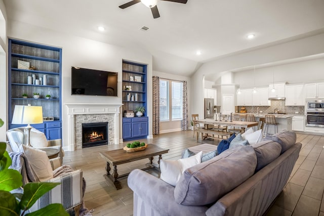 living room featuring a stone fireplace, vaulted ceiling, light hardwood / wood-style floors, and ceiling fan