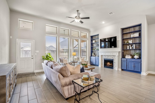 living room with ceiling fan, a stone fireplace, light hardwood / wood-style flooring, and built in features