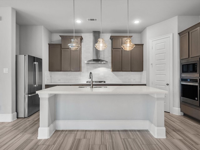 kitchen with stainless steel appliances, an island with sink, pendant lighting, and wall chimney exhaust hood