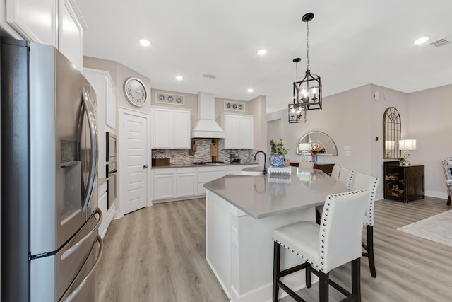 kitchen with premium range hood, appliances with stainless steel finishes, pendant lighting, white cabinetry, and a center island with sink