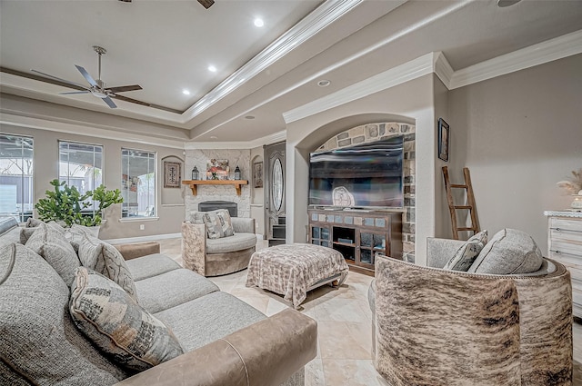 living room featuring a tray ceiling, a stone fireplace, ornamental molding, and ceiling fan