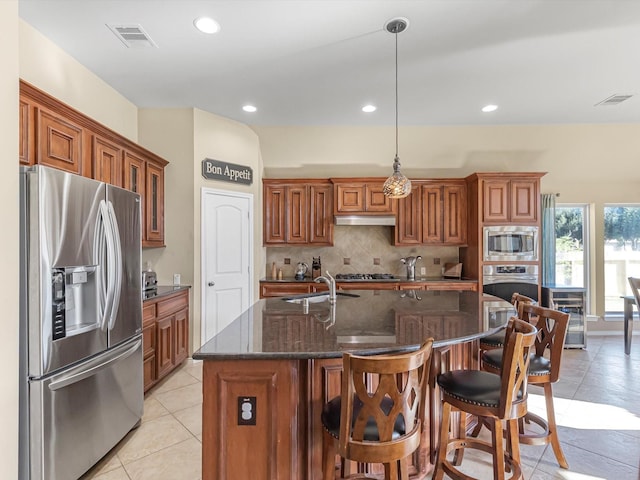 kitchen featuring decorative backsplash, hanging light fixtures, a kitchen island with sink, light tile patterned floors, and stainless steel appliances