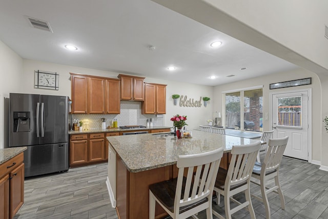 kitchen featuring stainless steel refrigerator with ice dispenser, tasteful backsplash, a kitchen breakfast bar, light stone countertops, and a kitchen island with sink