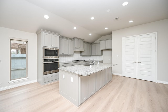 kitchen with lofted ceiling, appliances with stainless steel finishes, light stone counters, tasteful backsplash, and a center island with sink