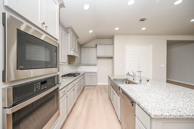 kitchen with sink, backsplash, stainless steel appliances, light stone counters, and a center island with sink