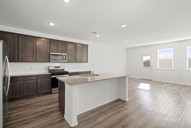 kitchen with hardwood / wood-style flooring, appliances with stainless steel finishes, light stone countertops, an island with sink, and decorative backsplash