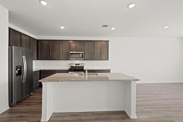 kitchen with appliances with stainless steel finishes, wood-type flooring, a kitchen island with sink, dark brown cabinetry, and light stone counters