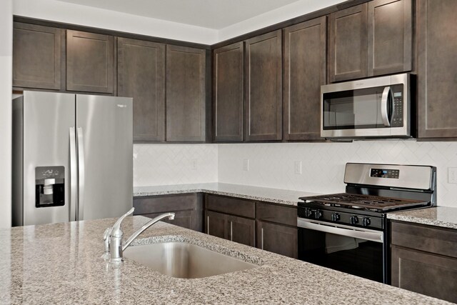 kitchen featuring sink, dark brown cabinets, stainless steel appliances, light stone countertops, and decorative backsplash