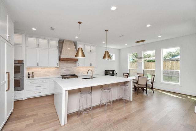 kitchen with sink, hanging light fixtures, an island with sink, custom range hood, and white cabinets