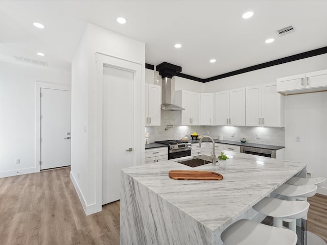 kitchen with stainless steel range with gas cooktop, white cabinetry, an island with sink, sink, and wall chimney exhaust hood