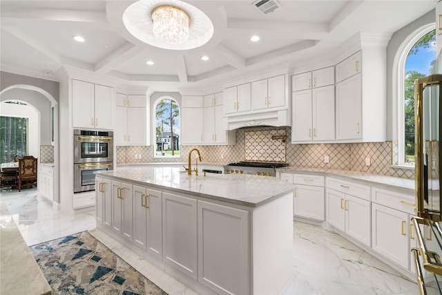 kitchen featuring a kitchen island with sink, stainless steel double oven, white cabinets, and light stone counters