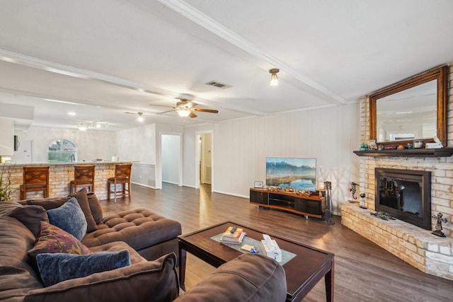 living room with a brick fireplace, beam ceiling, dark wood-type flooring, and ceiling fan