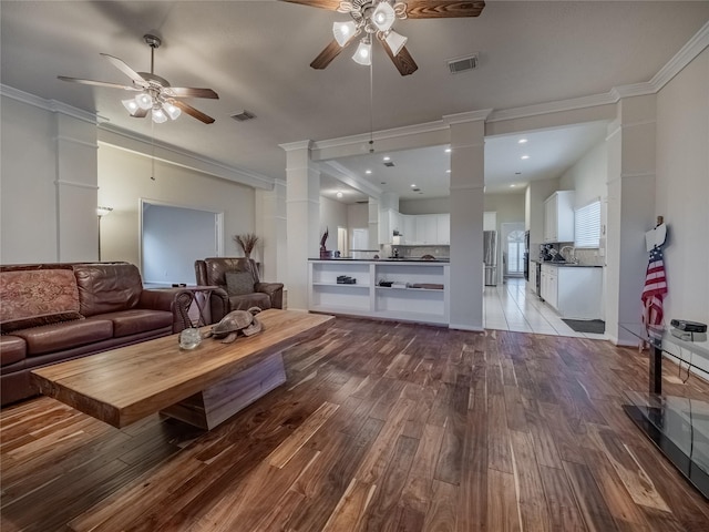 living room featuring ceiling fan, wood finished floors, visible vents, ornamental molding, and decorative columns