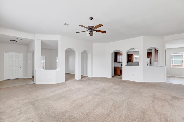 unfurnished living room featuring arched walkways, light colored carpet, visible vents, a ceiling fan, and baseboards