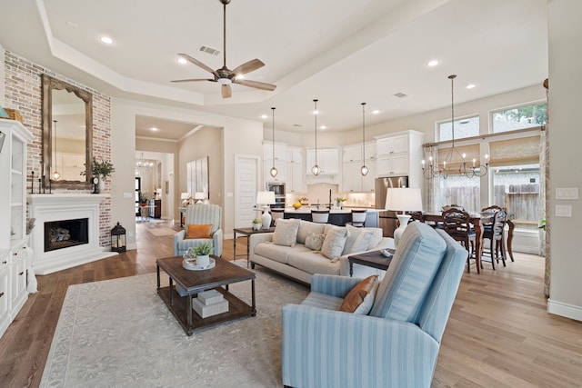 living room with a large fireplace, wood-type flooring, a raised ceiling, and ceiling fan with notable chandelier