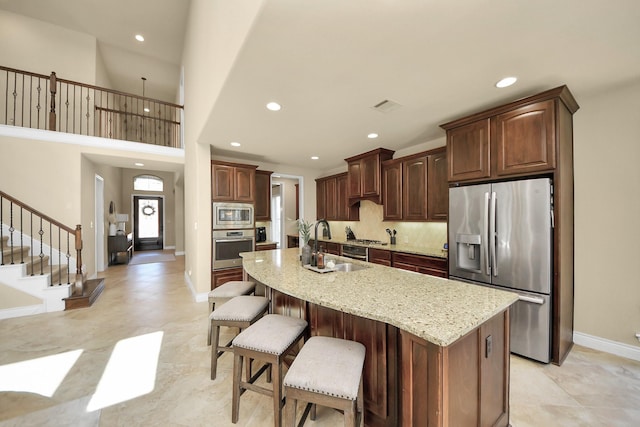 kitchen featuring a breakfast bar area, dark brown cabinets, a center island with sink, stainless steel appliances, and light stone countertops