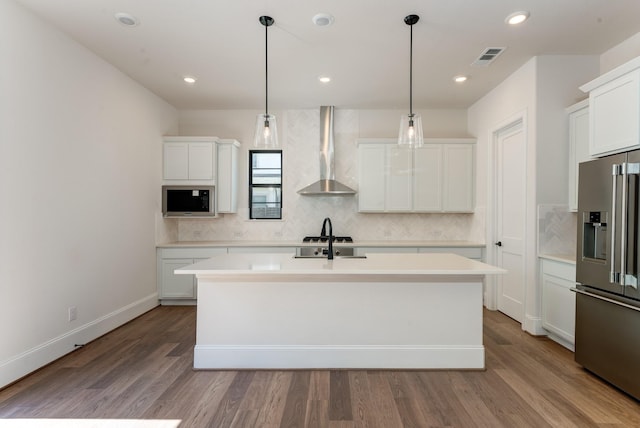 kitchen featuring decorative light fixtures, white cabinetry, wall chimney range hood, stainless steel appliances, and a center island with sink