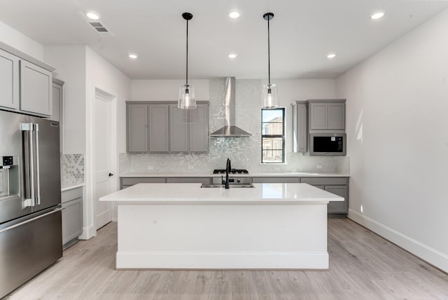 kitchen featuring sink, wall chimney range hood, hanging light fixtures, stainless steel appliances, and a center island with sink