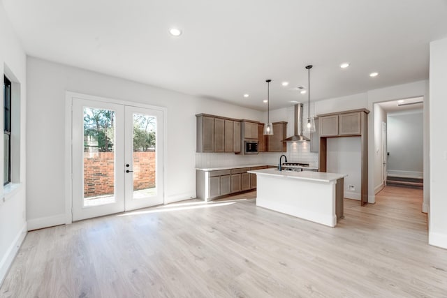 kitchen with wall chimney exhaust hood, hanging light fixtures, a center island with sink, light hardwood / wood-style floors, and backsplash