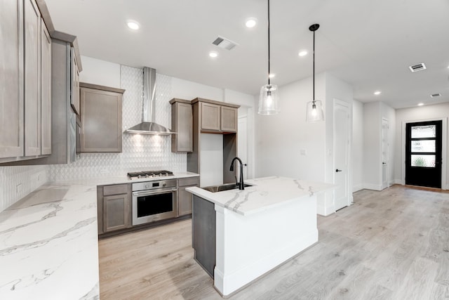 kitchen with wall chimney range hood, sink, appliances with stainless steel finishes, hanging light fixtures, and a center island with sink