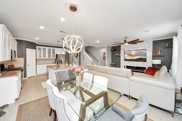 dining space featuring ceiling fan with notable chandelier, a fireplace, sink, ornamental molding, and light hardwood / wood-style flooring