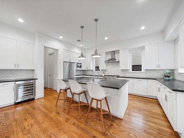 kitchen with high end refrigerator, dark countertops, wall chimney exhaust hood, wine cooler, and a kitchen bar