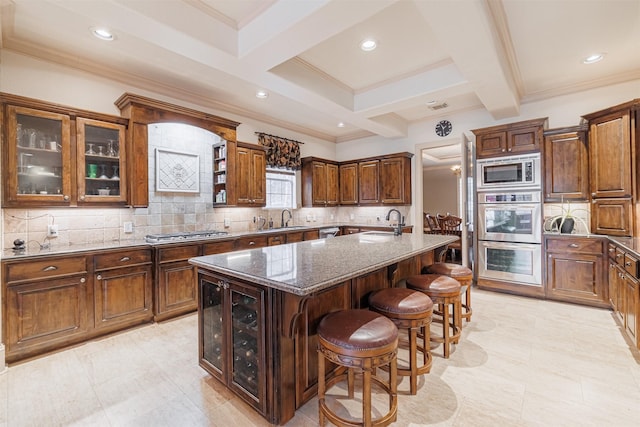 kitchen with beamed ceiling, sink, dark stone counters, stainless steel appliances, and a center island with sink