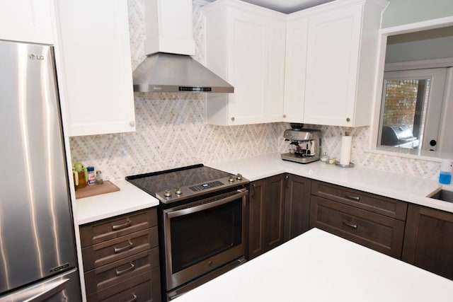 kitchen featuring appliances with stainless steel finishes, white cabinets, decorative backsplash, dark brown cabinets, and wall chimney range hood