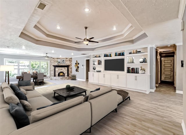 living room featuring a stone fireplace, crown molding, light wood-type flooring, a raised ceiling, and ceiling fan