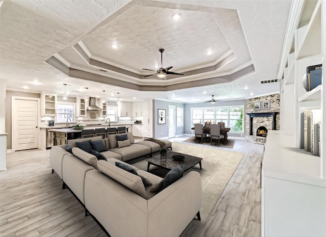 living room featuring a raised ceiling, ornamental molding, a stone fireplace, and light wood-type flooring