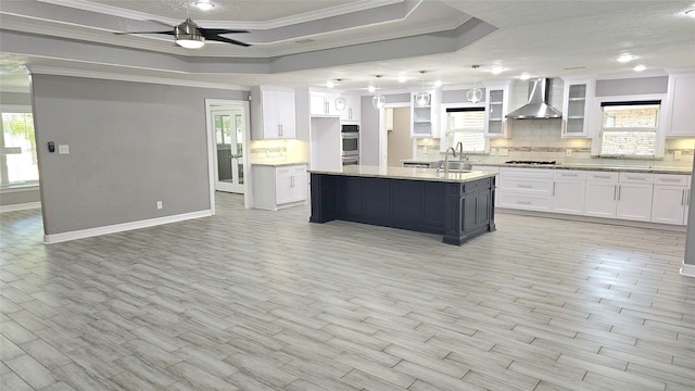 kitchen with a center island with sink, wall chimney range hood, white cabinets, and a tray ceiling