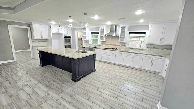 kitchen featuring white cabinetry, an island with sink, stainless steel double oven, gas stovetop, and wall chimney range hood