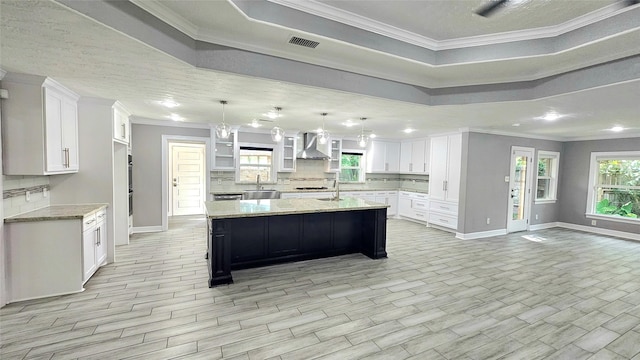 kitchen featuring wall chimney range hood, white cabinetry, hanging light fixtures, a spacious island, and light stone countertops