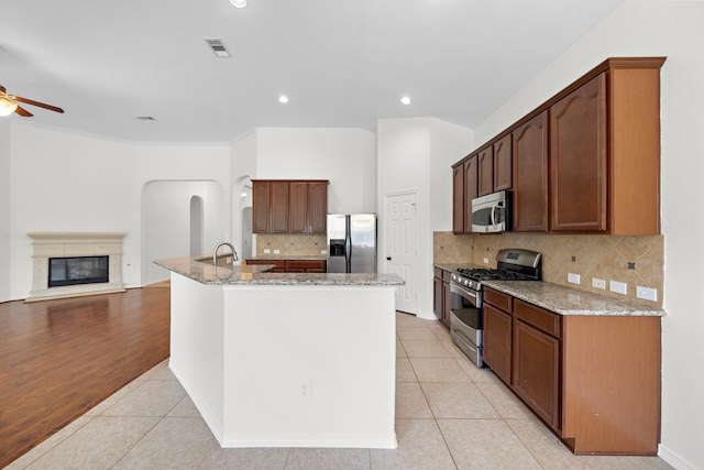 kitchen featuring ceiling fan, stainless steel appliances, light stone countertops, an island with sink, and light tile patterned flooring