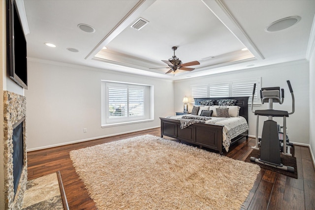 bedroom with a stone fireplace, dark hardwood / wood-style floors, ceiling fan, a tray ceiling, and crown molding