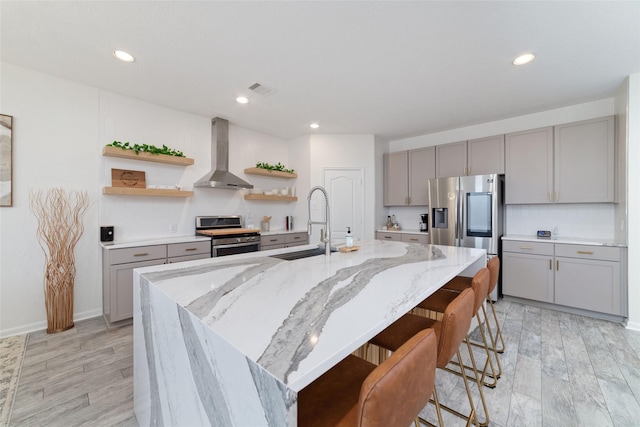 kitchen featuring wall chimney range hood, stainless steel appliances, a spacious island, light stone countertops, and a kitchen bar